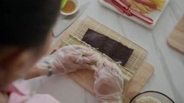 Nori 'yle kimbap yuvarlayan bir kızın ev mutfağında yemek pişirirken yüksek açılı görüntüsü.