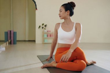 Genç Asyalı bir kadın yoga minderinde otururken rahat görünüyor ve minimalist stüdyoda ahşap döşeme ve raflarla odaklanıyor.