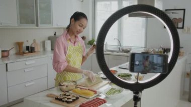 Z-Asyalı kızın ev mutfağında gimbap hazırlarken akıllı telefon ve halka ışığıyla video kaydettiği orta boy bir görüntü.