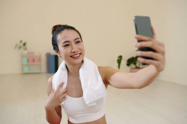 Spor salonunda selfie çekerken gülümseyen, spor kıyafeti giyen, akıllı telefon tutan, arka plandaki raflarda bitki ve kitaplarla minimalist bir odada duran bir kadın.