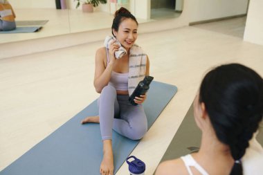 Yoga minderinde oturan, elinde su şişesi ve havluyla gülümseyen bir kadın. Parlak stüdyoda yoga seansından sonra sohbet ediyor. Aynaya yansıyan başka birinin oturup dinlediği görülebilir.