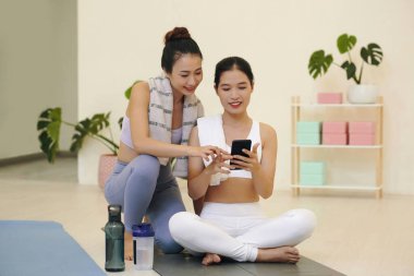 Yoga seansı sırasında egzersiz tekniklerini paylaşan iki genç Asyalı kadın, yoga minderlerine oturmuş su şişeleri ve yoga bloklarıyla birlikte, gülümsüyor ve rahatlıyor.
