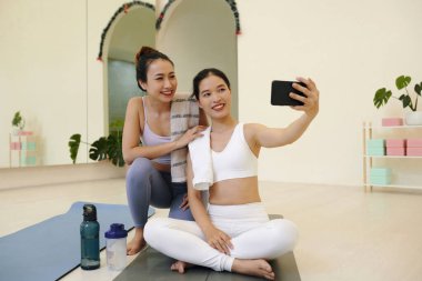 Yoga minderlerinde oturup selfie çeken iki genç kadın, modern stüdyoda bitkilerle mutlu bir şekilde gülümsüyor, rahatlama araçları görünür arka plan sakin bir ortam ve fitness rutinine odaklanmayı öneriyor.