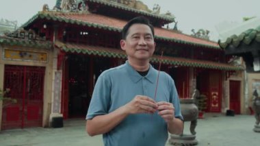 Antik tapınağı ziyaret ederken elinde yanan çubukla kameraya gülümseyen yetişkin Asyalı bir adamın orta boy portresi.