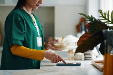 Yeşil üniformalı şahıs, arka planda tezgahta çeşitli mutfak malzemeleri ve malzemeleriyle modern mutfak ortamında sağlıklı yemek hazırlıyor.