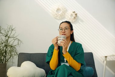 Hemşire kanepede oturuyor, elinde kahve fincanı tutuyor ve dikkatlice bakıyor. Arkaplan, pencereye yansıyan duvar dekoru ve güneş ışığını içeriyor.