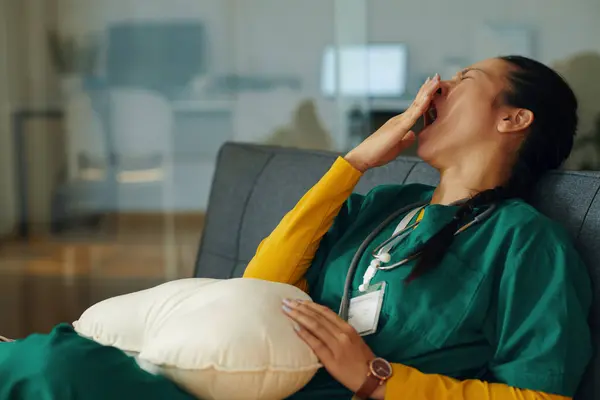 stock image Tired nurse in green scrubs yawning while resting during shift in dimly lit hospital room, wearing a stethoscope around neck