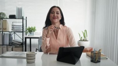 Tablet bilgisayarında çalışan beyaz iş kadınının orta boy portresi. Ofis masasındaki kameraya bakıyor.