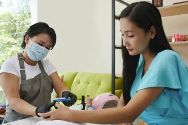 Servis sağlayıcısı, detaylara ve hijyen protokollerine odaklanmış, iyi ışıklandırılmış, çağdaş bir salon ortamında müşteriye manikür yapıyor.