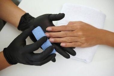 Siyah eldivenli bir spa uzmanının elleri müşteriye mavi tırnak törpüsü kullanarak manikür yaptırıyor, temiz, beyaz bir yüzeye dayanıyor ve dingin bir ortam yaratıyor.