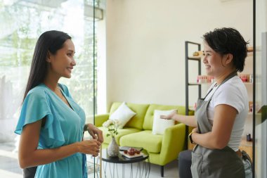 İki kadın parlak oturma odasında dikiliyor, biri önlük giyiyor. Samimi ve modern döşenmiş bir iç mekanda dostça bir sohbete giriyorlar.