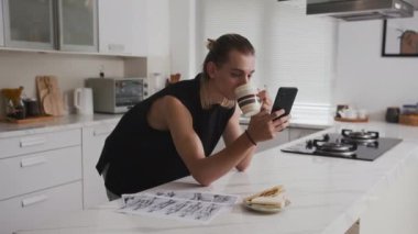 Genç beyaz erkek tasarımcı ev mutfağında kahvaltı yaparken cep telefonunu kontrol ediyor.