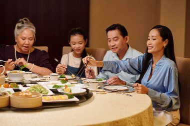 Asyalı bir aile restoranda yemek masasında oturuyor ve yemek yiyor.