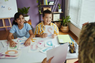 Eğitim materyalleri ve kitaplarla çevrili renkli mektuplarla etkileşimli bir sınıf aktivitesine katılan çocuklar, ön taraftan öğretmenler ise