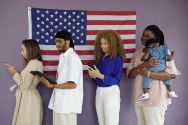 İnsanlar, oy verme merkezinde duvarında Amerikan bayrağı asılı dururken aletlerini kullanıyorlar.