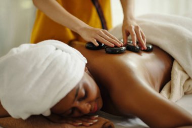 Spa merkezinde sıcak taş masajının keyfini çıkarmak, terapistin rahatlama terapisi için kadınlara sıcak taş atması, kafaya havlu sarılmış bir kadının rahatlaması.