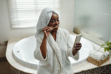 Dişleri beyaz bornozla fırçalayan, geniş banyoda akıllı telefon kullanan yuvarlak küvetli ve havluya sarılı pencereli biri.