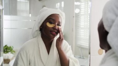 Beyaz bornozlu genç siyahi kadın evde güzellik hizmetleri sırasında göz altına takılan hidrojel bantlarını kontrol ediyor.