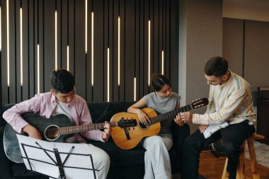 Modern bir müzik stüdyosunda takım çalışmasını ve yaratıcılığı vurgulayan samimi bir ambiyansı olan üç kişi birlikte gitar çalıyor. Doğrudan kameraya bakmıyorum.