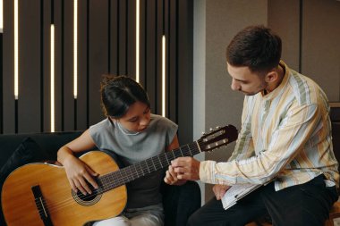 Adam, akustik gitarlı genç kıza akıl hocalığı yaparken şık bir odada kanepede oturuyor. Her ikisi de gitar ve defter ile öğrenme sürecine derinden bağlıydı.