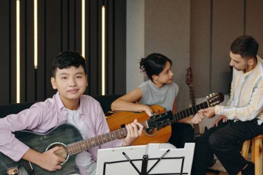 Gitar çalan üç kişinin portresi, biri kameraya bakıyor, diğeri akort yapıyor, ve üçüncüsü, rahat ve rahat bir şekilde oturuyor.