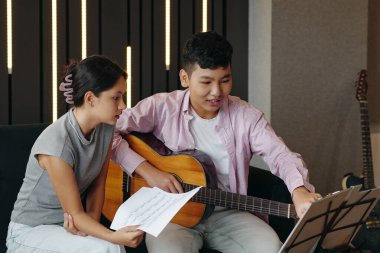 İki müzik meraklısı akustik gitar çalarken, arka planda dikey ışıklar ve gitarlarla modern stüdyoda otururken notalara odaklanıyorlar.