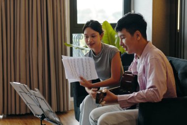 İçeride oturan iki kişi, gitar çalarak ve nota okuyarak müzikal bir projede işbirliği yapıyor, ilgi çekici ve üretken bir atmosfer yaratıyor.