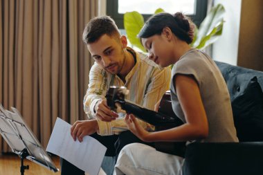 İki müzisyen kanepede oturup gitar çalarken ve müzik kağıtlarını incelerken, sıcak iç mekan ortamlarıyla çevrelenmiş, birlikte çalışmalar yapıyorlar.