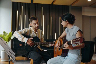 İki gitarist, müzik aletleriyle çevrili, iyi aydınlatılmış bir stüdyo ortamında teknikler ve ipuçları alışverişinde bulunurken pratik yapıyorlar.