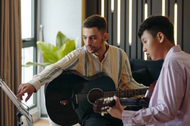 İki genç müzisyen birlikte akustik gitar çalıyorlar modern dekor ile rahat ev stüdyosunda, ayakta sergilenen notalar üzerinde yoğunlaşıyorlar, pencereden içeri güneş ışığı giriyor.