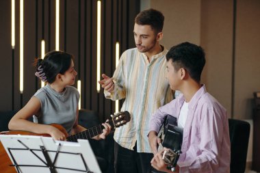 Modern iç mekanda akustik gitarlar ve notalarla müzik pratiği yapan bir grup yetişkin. Gitar tekniğine yardımcı olan eğitmen.