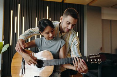 Modern tasarım oturma odasında yetişkin bir adamın yardımıyla gitar öğrenen genç bir kız. Bir adam gülümseyerek çocuklarına rehberlik ederken gitarın üzerine elini koyuyor.