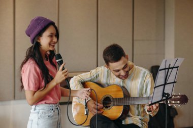 Genç Asyalı kadın, müzik stüdyosunda gitar çalan beyaz adamın yanında mikrofonla şarkı söylüyor, gülümsüyor ve müzikal işbirliğinden zevk alıyor.