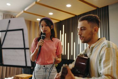 Elinde mikrofon tutarken şarkı söyleyen genç bir kadın, bir müzik seansı sırasında bir akustik gitarist eşliğinde arka planda aydınlanma olan bir stüdyoda.