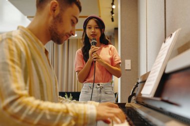 Genç Asyalı kadın mikrofonla şarkı söylerken, modern stüdyoda piyano çalan genç çift ırklı adam müzik performansına odaklanıyor.