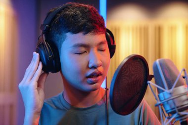 Genç Asyalı erkek, ses geçirmez stüdyoda şarkı söylerken tek eliyle kulaklık tutuyor ve mikrofonun önünde dikiliyordu.