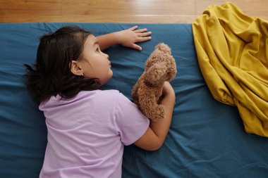 Oyuncak ayısıyla yatakta dinlenen küçük kızın yüksek açılı görüntüsü