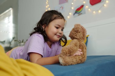 Küçük kız odada yatağında oyuncak ayıyla oynuyor.