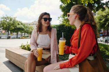 İki genç kadın ellerinde portakal suyu, güneşli bir parkta ahşap bir bankta otururken sohbet ediyorlar.