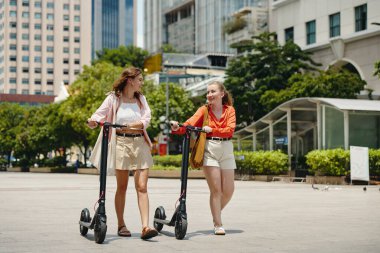 İki arkadaş, binalar ve yeşilliklerle çevrili kentsel bir ortamda, her biri elektrikli scooterla sohbet ederken, dışarıda gezinmenin keyfini çıkarıyorlar.