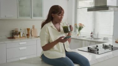 Genç siyahi bir kızın yeşil smoothie içerken ve akıllı telefonunu kontrol ederken sabahını evde mutfakta geçirdiği orta boy bir fotoğraf.