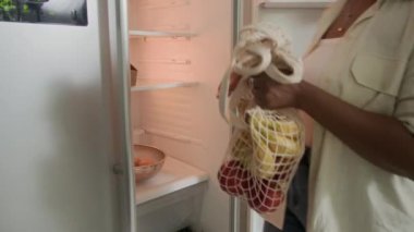Mutfaktaki buzdolabına ağ torbalarını boşaltan bir kadın resmi.