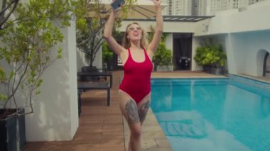 Mayo ve güneş gözlüklü neşeli beyaz kızın tatilde havuz kenarında akıllı telefon kullanarak dans edişinin fotoğrafını çekiyorum.