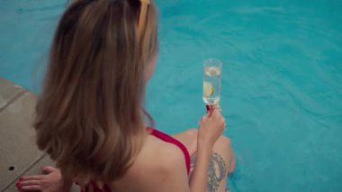 Yaz tatili boyunca taze limonlu içecekle rahatlarken bacaklarını yüzme havuzuna daldıran kırmızı mayo giymiş genç, beyaz bir kadın.