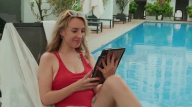 Yaz tatilinde havuz başında dinlenirken dijital tablet kullanan genç beyaz kadının el kamerası görüntüsü.