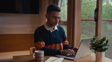 Bilgisayara e-posta yazan ve ofis masasında otururken kameraya gülümseyen pozitif erkek serbest çalışanın orta boy portresi.