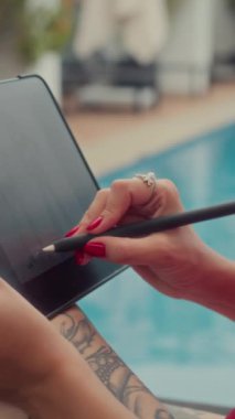 Tatil köyünde havuz başında dinlenirken dijital tablete çizim yapan isimsiz turistlerin dikey çekimleri