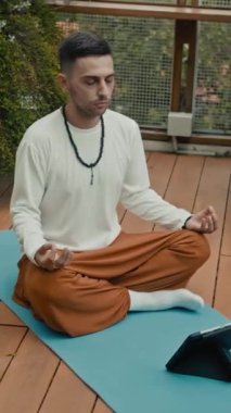 Genç adamın lotus asana 'da tabletin önünde nefes egzersizi yaparken dikey çekimi.