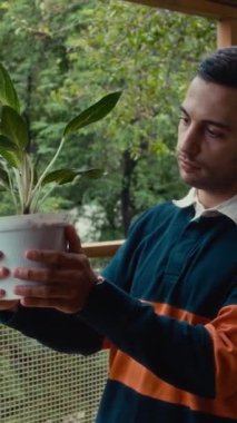 Evdeki balkonda elinde çiçekle gülümseyen bir gencin dikey portresi.