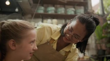 Neşeli Asyalı bayan öğretmenin çömlekçide çömlek tekeri üzerinde kil çömlek ustası genç beyaz kızı eğip...